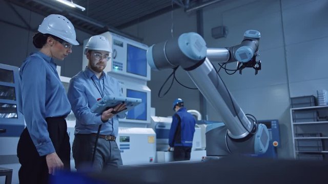 Factory Workshop: Professional Female Engineer, Male Machinery Operator Use Industrial Digital Tablet Computer to Work and Program Robot Arm for Production Line. High-Tech Facility with CNC Machines