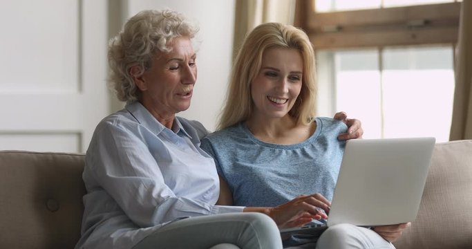 Pleasant elderly middle aged woman sitting on couch embracing grown up daughter, watching family or journey photos on computer. Smiling blonde lady listening to older nature mommy, using laptop.