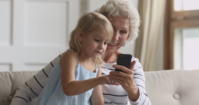 Interested little preschool child girl using mobile phone with smiling elderly grandmother, sitting together on cozy sofa. Happy middle aged granny holding smartphone, while small kid playing on it.