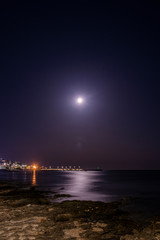 The full moon is reflected on the water of the sea, in a beautiful night landscape. In Italy, Puglia, Lecce, Salento. The marina with boats moored in the background. The rocks in the foreground.