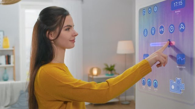 Beautiful Young Female Interacts with Augmented Reality Display. She's Changing Climate Control Setting to Higher Temperature. Smart Home Concept with Transparent Screen Made with VFX Special Effects.