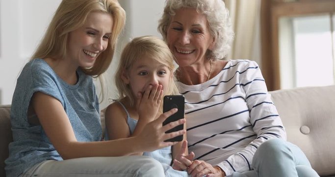 Happy young woman sitting on couch with excited older mother and little preschool daughter, using funny photo editing applications on smartphone. Joyful family recording short videos together at home.