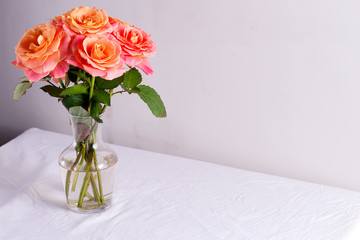 Glass vase with beautiful bouquet of fresh natural roses flowers on a table covered white cloth against light grey wall.