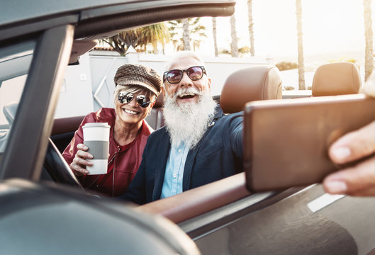 Happy senior couple taking selfie on new modern convertible car - Mature people having fun together making self photos during road trip vacation - Elderly lifestyle and travel transportation concept
