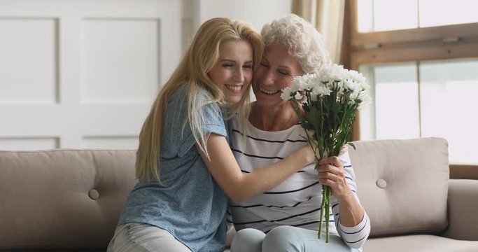 Smiling young blonde woman giving flowers bouquet to happy middle aged older mother, congratulating with birthday, international woman s day or special occasion, sitting together on sofa at home.