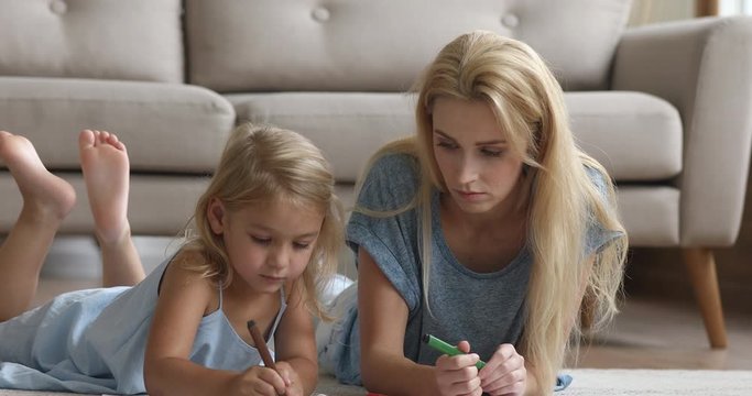 Happy young attractive woman lying on light carpet with adorable small preschool daughter, drawing pictures with colorful markers. Millennial female babysitter teaching little girl writing letters.