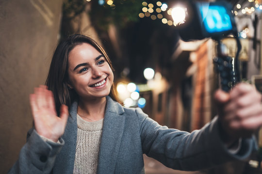 Female blogger showing hi recording video for vlog social media with digital camera. Smiling woman vlogger taking photo selfie on background illuminations light night city