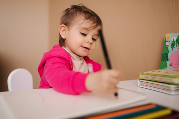 Naklejka premium Happy baby girl sit at the table and wthire something. Little girl use pencil for drawing on white paper at home