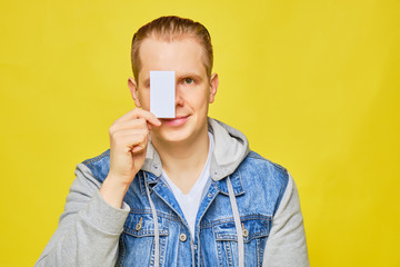 Stylish Caucasian man in a jeans on a yellow background holds a white card at eye level and smiles copy space.