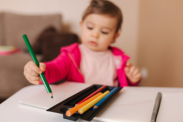 Happy baby girl sit at the table and wthire something. Little girl use pencil for drawing on white paper at home