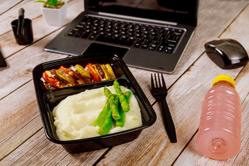 Lunch box container with mashed potato, asparagus and vegetables on wooden table with laptop.