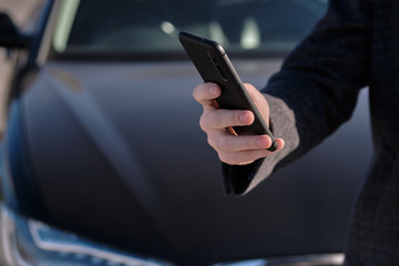 A man in a coat holds a smartphone on the background of the car.