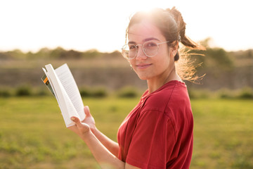 Young woman having a nice reading while smiling and enjoying the light of a sunset in a park.