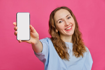 Stylish caucasian girl in a pale blue t-shirt holds a smartphone in his hand and shows it to the camera on a pink background. Copy space.