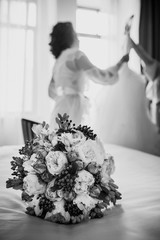 Elegant wedding white dress hanging on a wooden window during a wedding preparation. Bride's morning. Wedding bouquet on a bed. Before ceremony