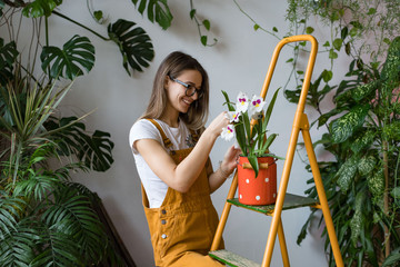 Young smiling woman gardener in glasses wearing overalls, taking care for orchid in old red milk...