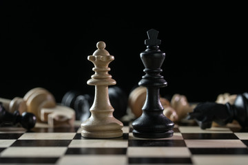 Chess pieces queen and king in front of a of fallen chessmen after a battlefield on a chessboard against a black background, concept of abuse of power, civil victims and the lack of responsibility