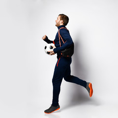 Soccer player in blue tracksuit, sneakers and backpack. Jumping up, smiling, holding ball, posing isolated on white. Full-length