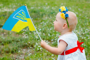 little girl with Ukrainian embroidery and wreath with yellow and blue flowers is carried by Ukrainian flag. Happy baby kid on Independence Day of Ukraine and the flag day