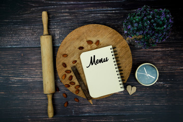 Menu on notebook with boards, pencil, roller pin, love origami, clock, flower and beans. Top view.