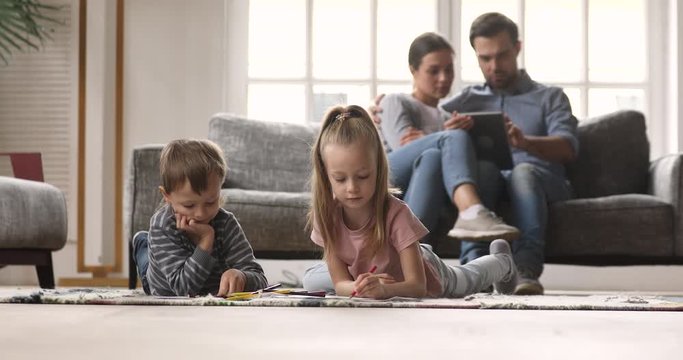 Cute small brother and sister lying on floor carpet, drawing coloring pictures in paper album while young loving couple parents relaxing on comfortable sofa, using digital computer tablet at home.