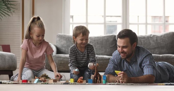 Happy little children constructing building with wooden blocks with young father, lying together on warm floor. Smiling daddy enjoying weekend leisure playtime with small cute kids in living room.
