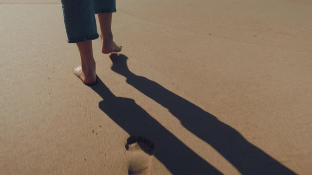 Low angle, slow motion and close up shot of an unrecognisable woman walking barefoot in a beautiful summer vibe beach. A girl is wearing blue jeans.