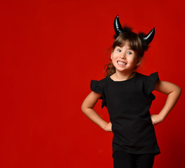 Little girl with devil horns, in black blouse and leggings. She laughing, hands on hips, posing on red background. Close up