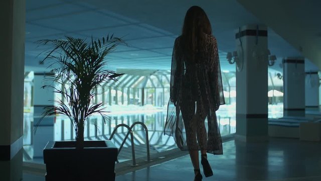 The young girl goes down the stairs towards the hotel's glass-covered indoor pool. she takes off her pareo and stays in her bikini. back view. slow motion. low light