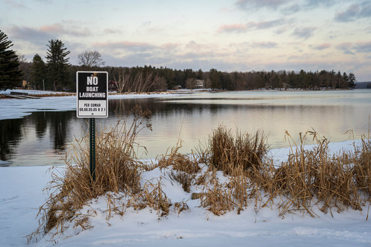 A metal sign marking a no-boat launch area, stands in the ground on the shore of a half frozen lake covered in ice and snow.