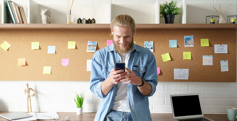 Smiling millennial hipster man standing at home office workspace using cell phone...