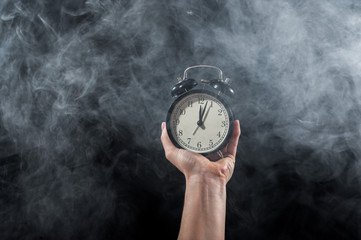Close-up of a female hand holding a clock on a black background in smoke. Alarm clock at midnight in a mystical fog.