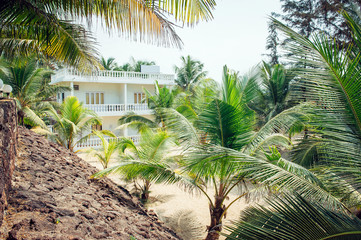 Fototapeta na wymiar View of palm trees on the sand and a white stone building with balconies. Small hotel, guesthouse in Asia. Sunny tropical afternoon.