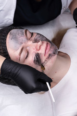 Application of cosmetology mask on face of young woman. Acne treatment. Black carbonated mask. Procedure for face skin rejuvenation. Spa, cosmetology and preservation of youth. Wellness concept.