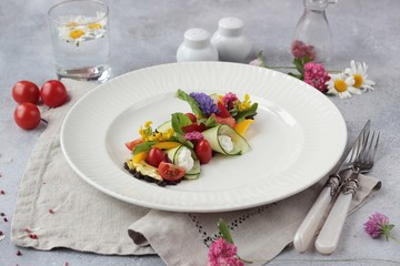 Colorful vegetable salad decorated with flowers on a white plate, light grey stone background. Healthy spring or summer lunch.