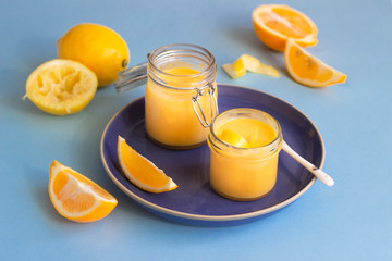 Two jars of homemade lemon curd or custard and fresh lemons on a blue ceramic plate. Blue paper background. Selective focus.