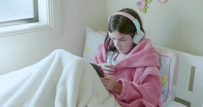 a young girl sick in bed watching a tablet computer, with headphones on, she is wearing a pink robe, there is a box of tissues next to her, the camera pans around her slowly. She is in isolation