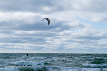 A person in a special black suit engaged in kitesurfing moves to the right behind the kite in the distance on the horizon of an unsettling stormy blue sea and cloudy overcast sky on a windy day.
