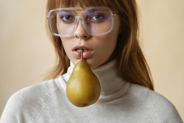 Fashion young lady hold pear in mouth look at camera, stylish girl pretty face wear trendy glasses posing, vogue sexy attractive model in eyewear on beige studio background, close up view portrait
