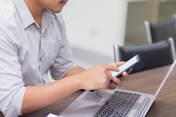 A man's hand is pressing on the smartphone screen on the table and the laptop is in the background. A business man eating coffee in a paper cup and pressing the phone with white screen.