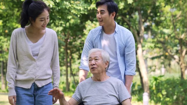 young asian couple taking senior father for a walk and helping him stand up from wheel chair