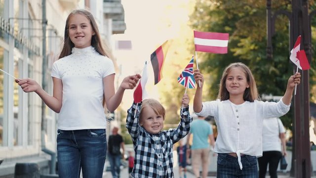 Group of kids holding different flags. Children are in city center. Pupils have Canadian, German, Czech, Austrian, British, and Indian flag.