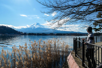 Man leaning against the bridge over Kawaguchiko Lake, Japan with the view on Mt Fuji. The mountain is surrounded by clouds. Man is enjoying the walk and good weather. Serenity and calmness