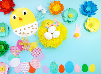 Child made crafts his own hands at Easter. Handmade nest with eggs, flowers, chicken, chicken in an egg, frame of eggs. Cute art creativity on blue background. Flat lay, top view, close up, copy space