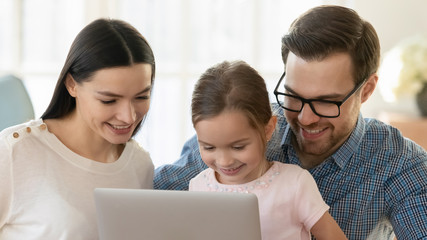 Head shot close up happy married couple relaxing with little daughter, using computer together at home. Smiling parents watching funny videos cartoons with kid girl, shopping making purchases online.