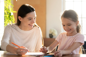 Head shot young happy babysitter tutor educating little preschool child girl, sitting together at...