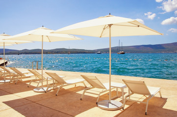 Summer beach vacation. Lounge chairs  and white sun umbrellas against blue sky. Montenegro, Adriatic Sea, Bay of Kotor, Tivat city