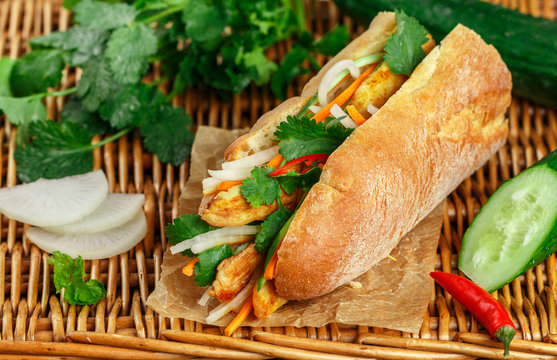 Baguette sandwich with fried chicken and fresh vegetables-carrots, cucumber, daikon, red pepper and cilantro (coriander) on a wicker table. Delicious fast food. Vietnamese Banh Mi. Selective focus