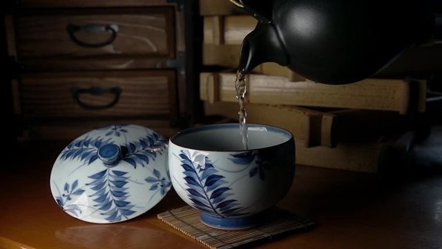 Light Japanese tea is poured from a dark brown clay pot into an old chipped tepid white porcelain cup