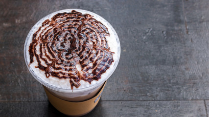 Iced latte with milk foam and chocolate sauce on top, in a plastic cup on wooden table background, and copy space.
